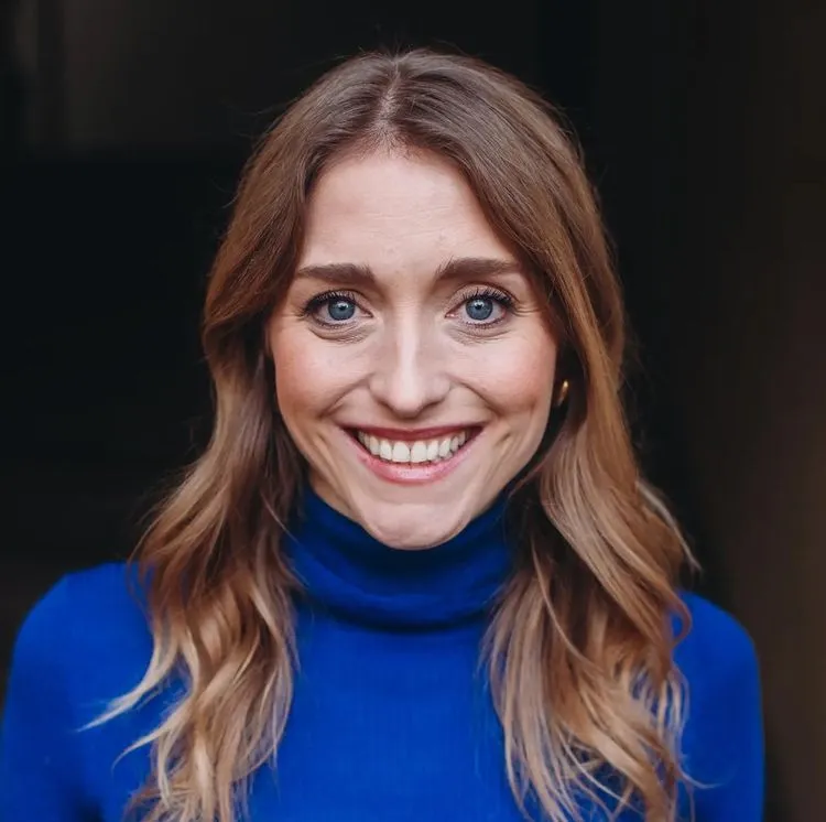 Smiling woman with long light-brown hair wearing a bright blue turtleneck, facing the camera against a dark, softly blurred background.