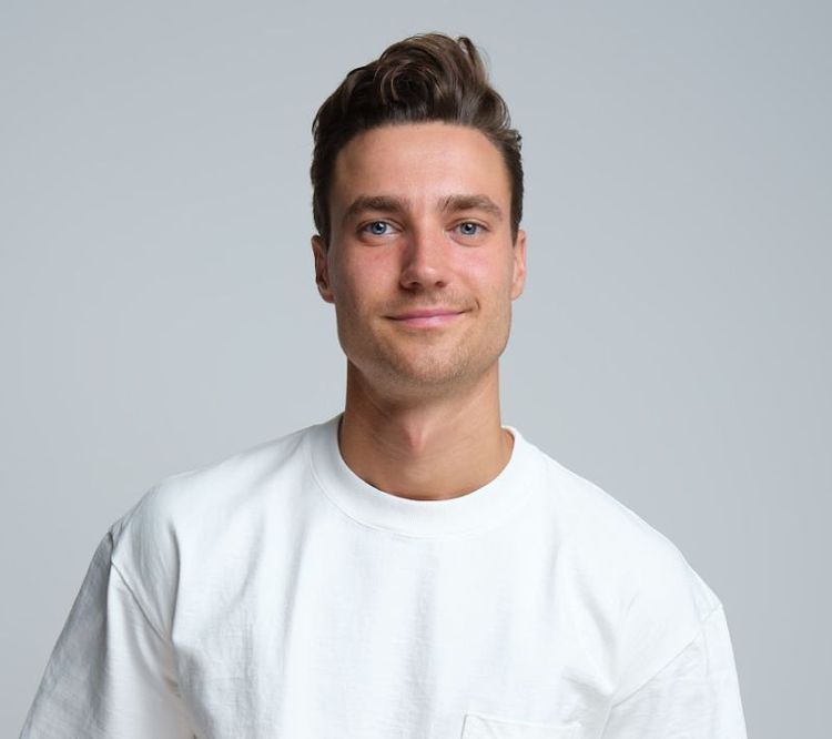 Man with short brown hair and light stubble wearing a plain white T-shirt, looking directly at the camera against a light gray background.