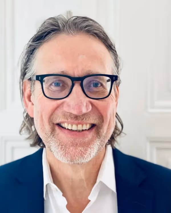 Smiling man with glasses and short graying hair, wearing a navy blazer and white shirt, seated at a table with hands clasped in a modern indoor setting with plants and wooden elements in the background.