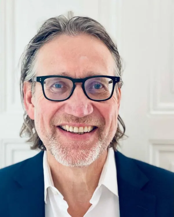 Smiling man with glasses and short graying hair, wearing a navy blazer and white shirt, seated at a table with hands clasped in a modern indoor setting with plants and wooden elements in the background.