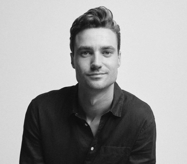 Man with short brown hair and light stubble wearing a plain white T-shirt, looking directly at the camera against a light gray background.