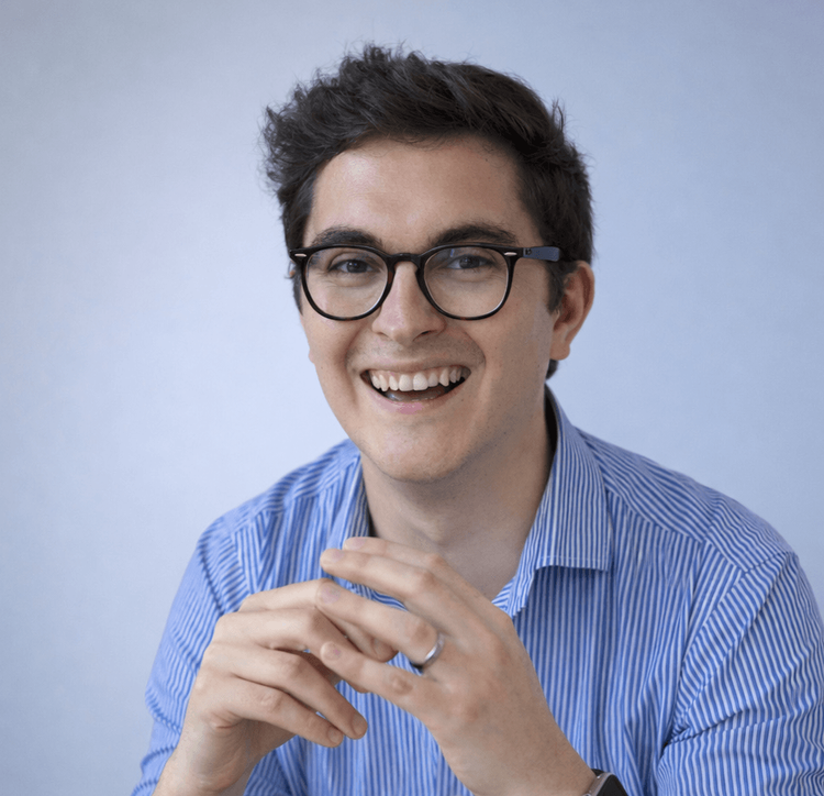 Portrait of Gabi Lipan smiling, wearing glasses and a blue striped shirt, photographed against a clean light background.