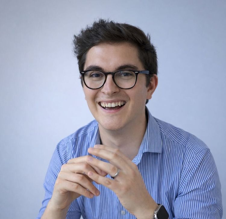 Portrait of Gabi Lipan smiling, wearing glasses and a blue striped shirt, photographed against a clean light background.