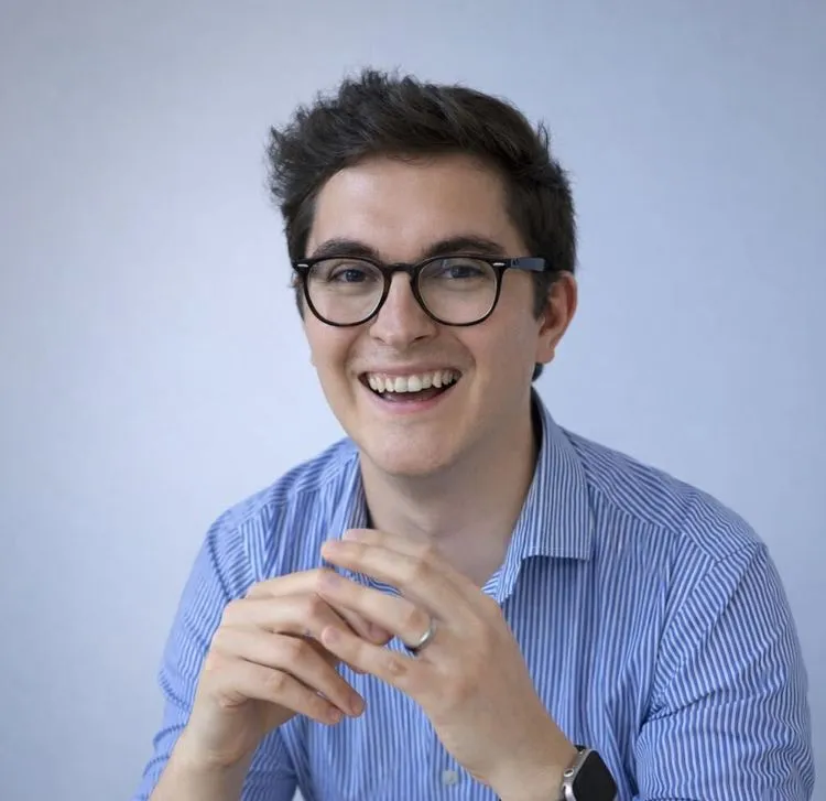 Portrait of Gabi Lipan smiling, wearing glasses and a blue striped shirt, photographed against a clean light background.
