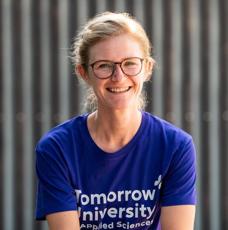 Portrait of Maren Kropfeld smiling, wearing glasses and a blue “Tomorrow University of Applied Sciences” T-shirt, photographed outdoors against a vertical metal background.