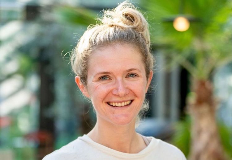 Portrait of Maren Kropfeld smiling outdoors, wearing a light-colored top with hair tied up, photographed in a bright, green, plant-filled setting.
