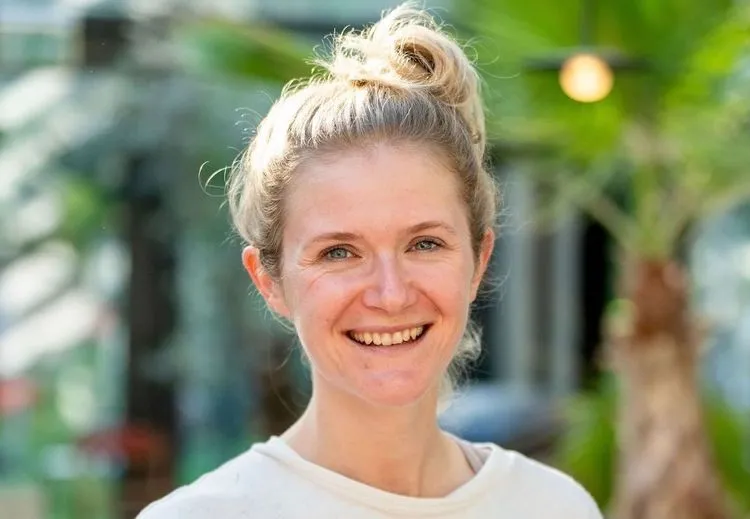 Portrait of Maren Kropfeld smiling outdoors, wearing a light-colored top with hair tied up, photographed in a bright, green, plant-filled setting.