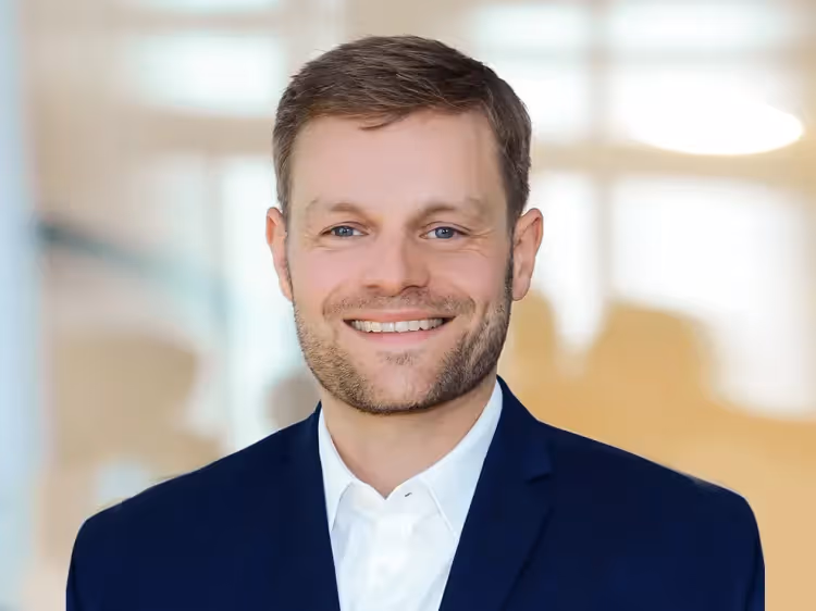 Portrait of Matthias Lange smiling, wearing a dark suit and white shirt, photographed against a softly blurred, light-filled office background.