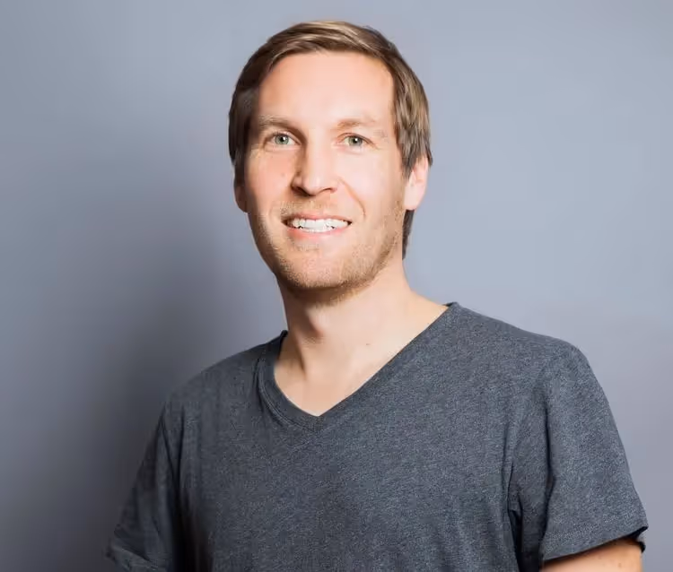 Portrait of Tim Schumacher smiling, wearing a gray T-shirt, photographed against a neutral gray background.