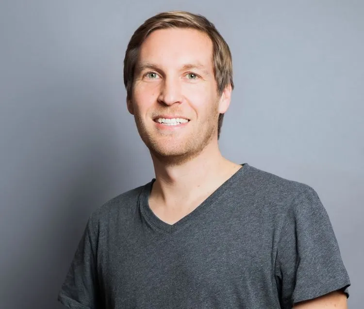 Portrait of Tim Schumacher smiling, wearing a gray T-shirt, photographed against a neutral gray background.