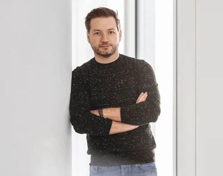 Portrait of Rolf Schrömgens standing with arms crossed, wearing a dark sweater and jeans, photographed in a bright, modern interior with soft natural light.
