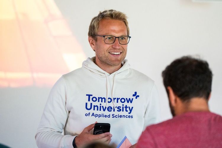 Thomas Funke wearing glasses and a white ‘Tomorrow University of Applied Sciences’ hoodie, holding a phone while speaking to another person in an indoor setting.