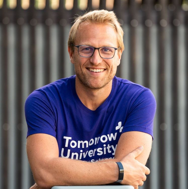 Thomas Funke wearing glasses and a blue ‘Tomorrow University’ T-shirt, leaning forward with arms crossed against a blurred outdoor background.