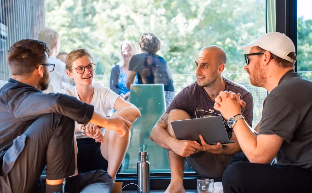 Small group of people sitting together by a window, talking and collaborating with a laptop in a bright indoor space.