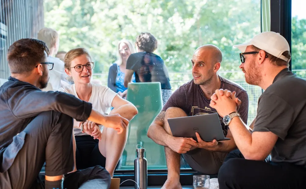 Small group of people sitting together by a window, talking and collaborating with a laptop in a bright indoor space.