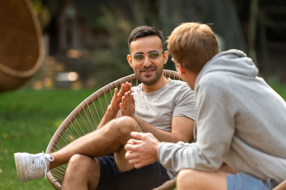Two people sitting outdoors on chairs in a green space, engaged in a relaxed conversation.