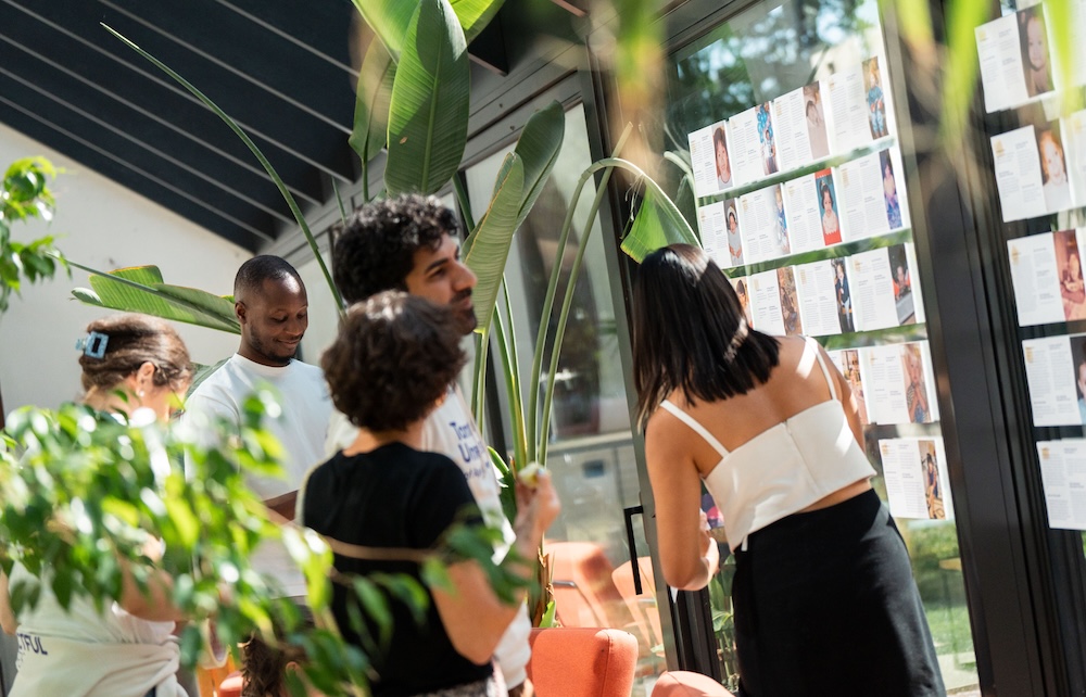 Group of people standing together indoors, looking at a wall display with printed profiles surrounded by plants.