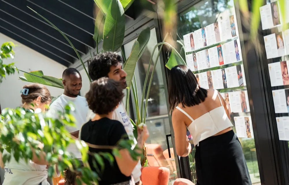 Group of people standing together indoors, looking at a wall display with printed profiles surrounded by plants.