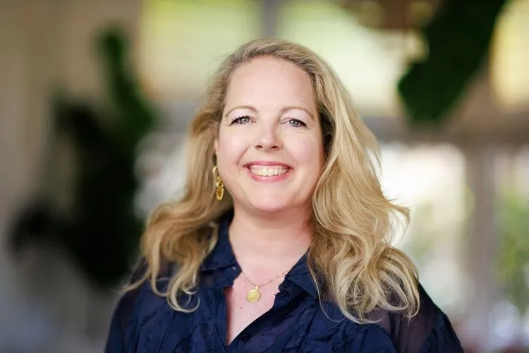 Smiling woman with long blonde hair wearing a dark blouse and gold jewelry, standing indoors with soft natural light and blurred green plants in the background.