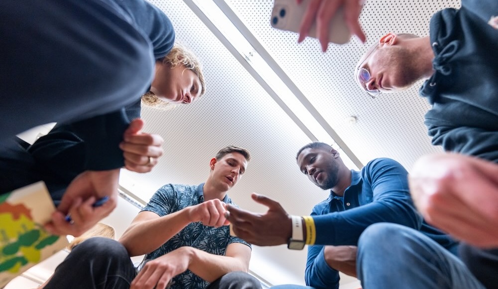 Low-angle view of a diverse group of young adults sitting in a circle indoors, leaning in and discussing something with papers and a smartphone in hand.