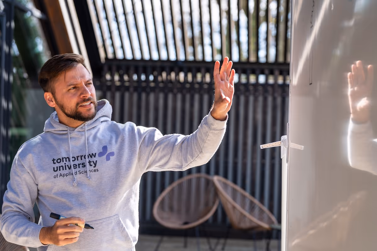 A man wearing a grey Tomorrow University of Applied Sciences hoodie holding a marker and gesturing toward a whiteboard in a bright outdoor setting.