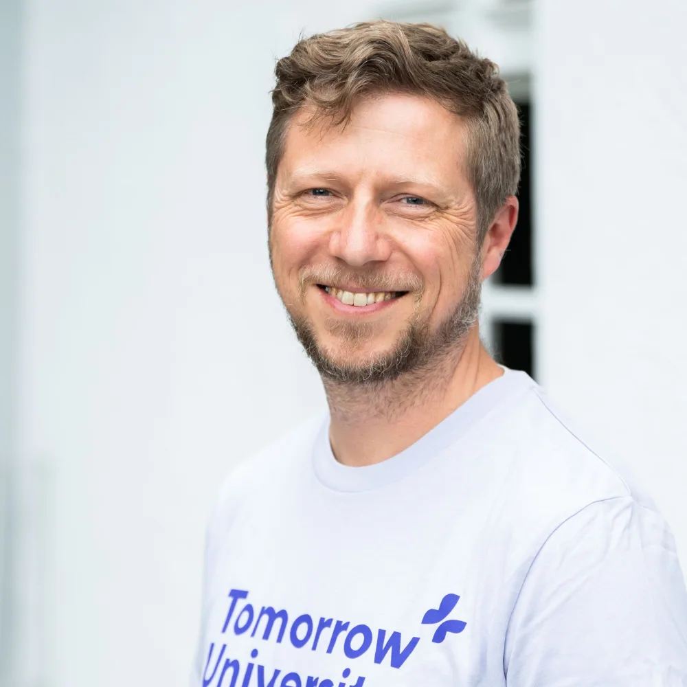 Portrait of Andrew Elliott smiling, wearing a light lavender Tomorrow University T-shirt, photographed against a bright white wall.