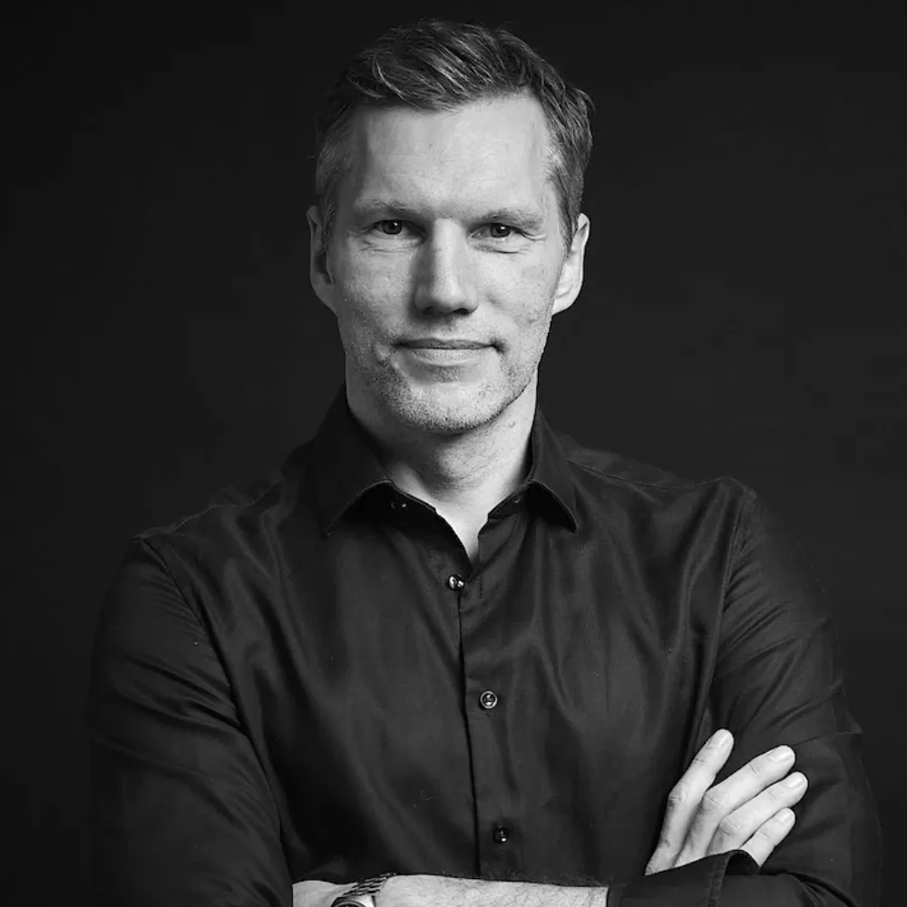 Black-and-white portrait of Benjamin Körner, arms crossed, wearing a dark button-down shirt, photographed against a dark background.