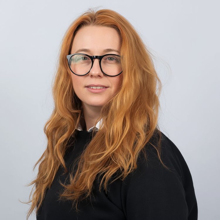 Professionelles Porträt von Alisa Reznik mit langen roten Haaren und Brille, in weißem Kragenhemd und schwarzem Pullover, sanft lächelnd vor hellgrauem Hintergrund.