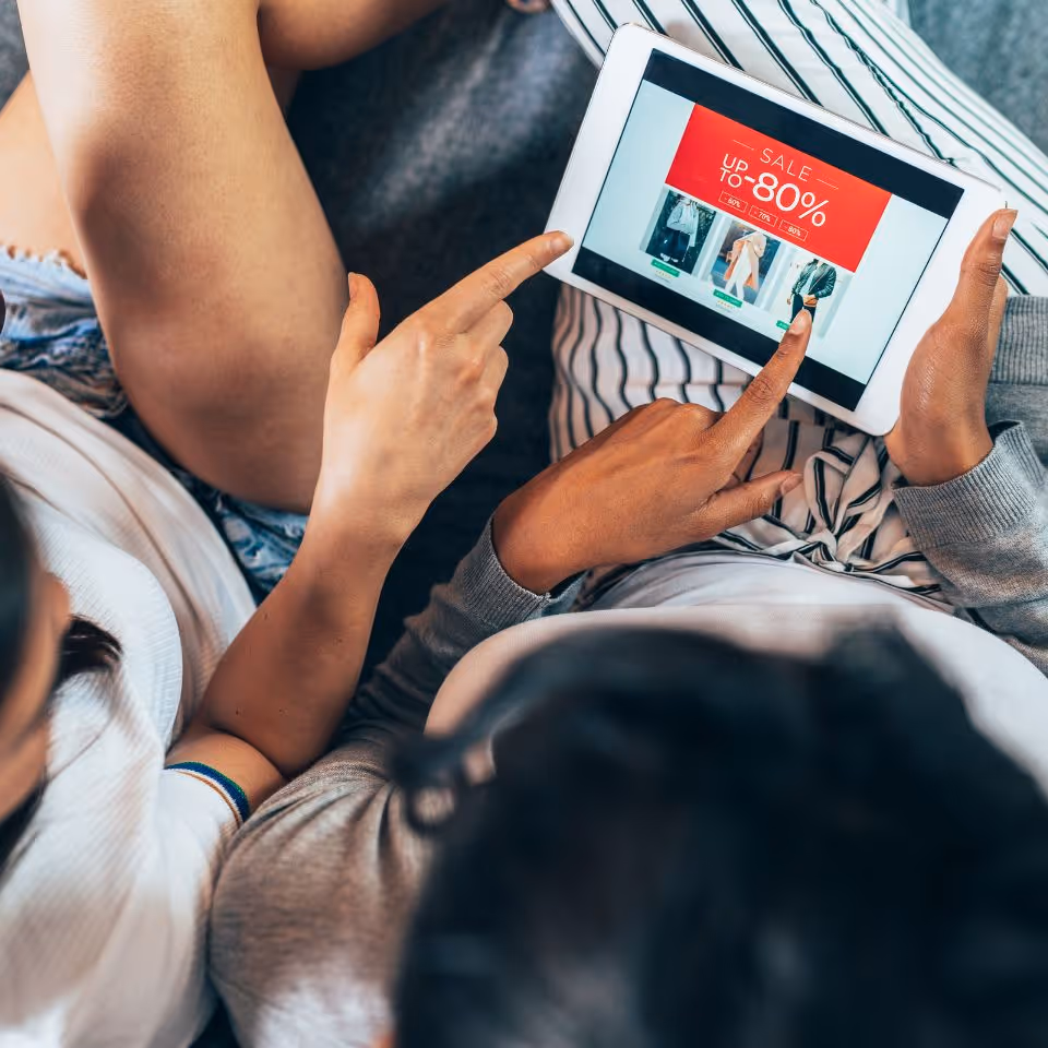 Two people sitting close together browsing a tablet showing a sale up to 80% off on clothing items.