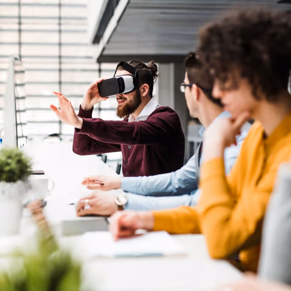 Man using virtual reality headset at a desk with colleagues in a modern office.