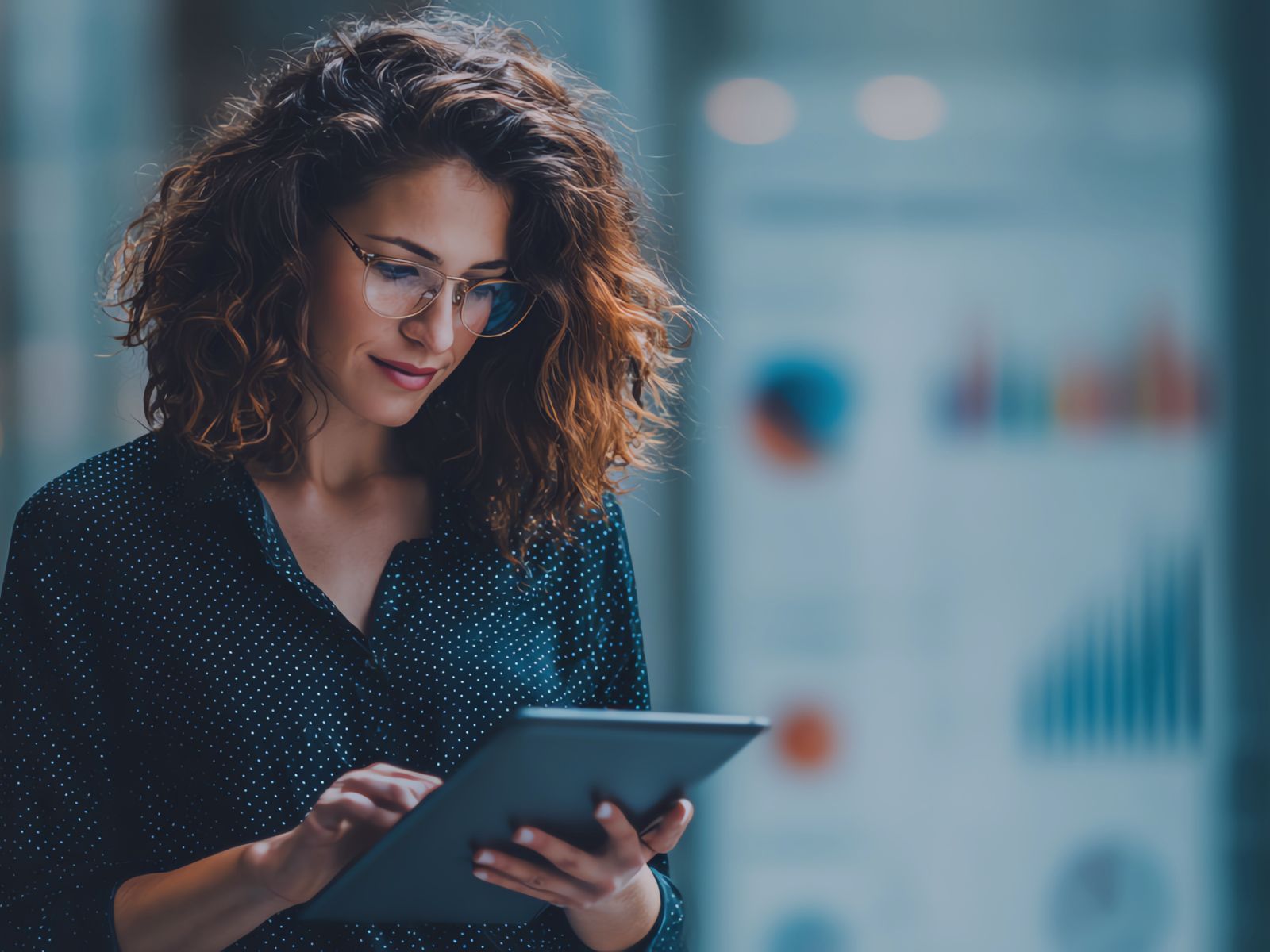 Woman with curly hair and glasses using a tablet with blurred charts in background.