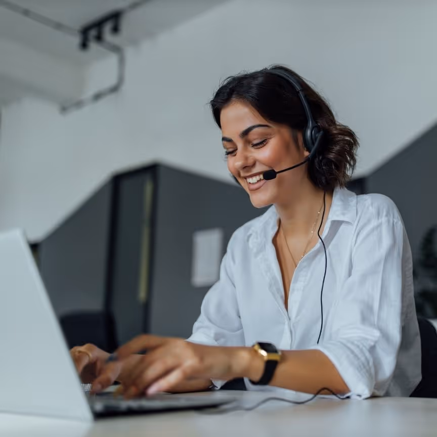 Smiling woman wearing headset typing on laptop while making video call at modern office desk.