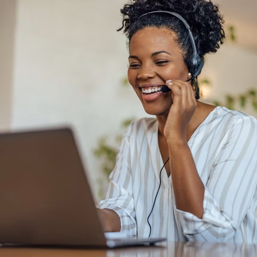 Smiling woman wearing headset, talking and working on laptop, likely customer support agent.