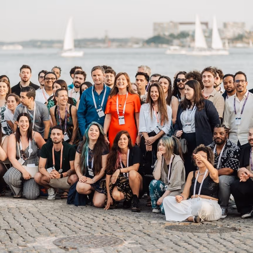 Large diverse group of people posing outdoors by waterfront with sailboats in background.