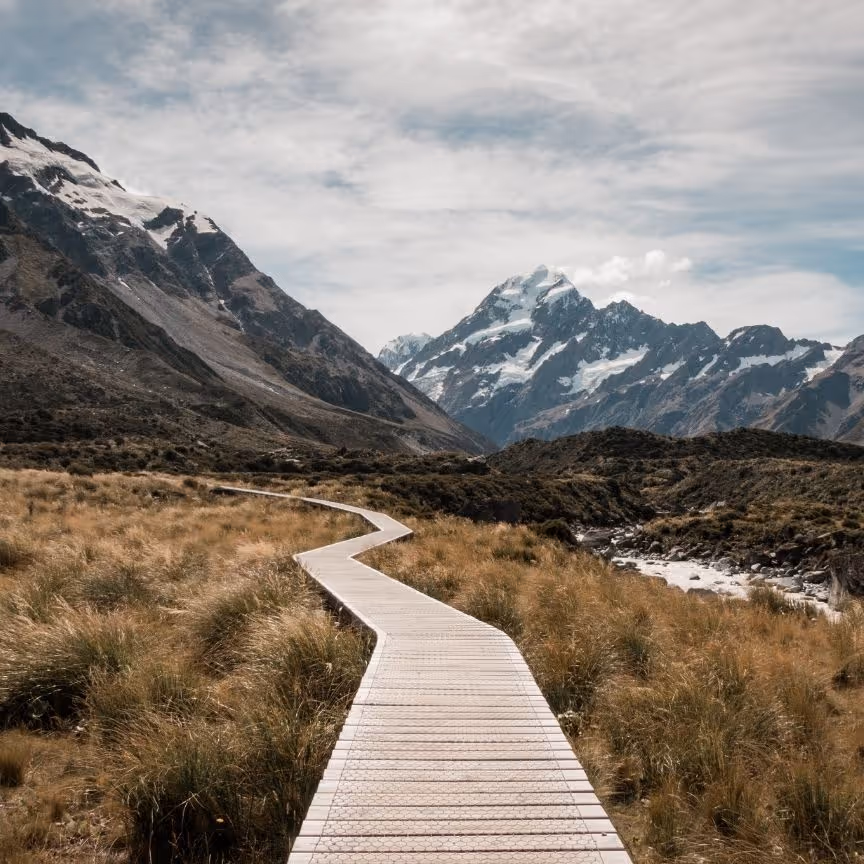 Wooden path winding through grassy terrain toward snow-capped mountains under a cloudy sky.