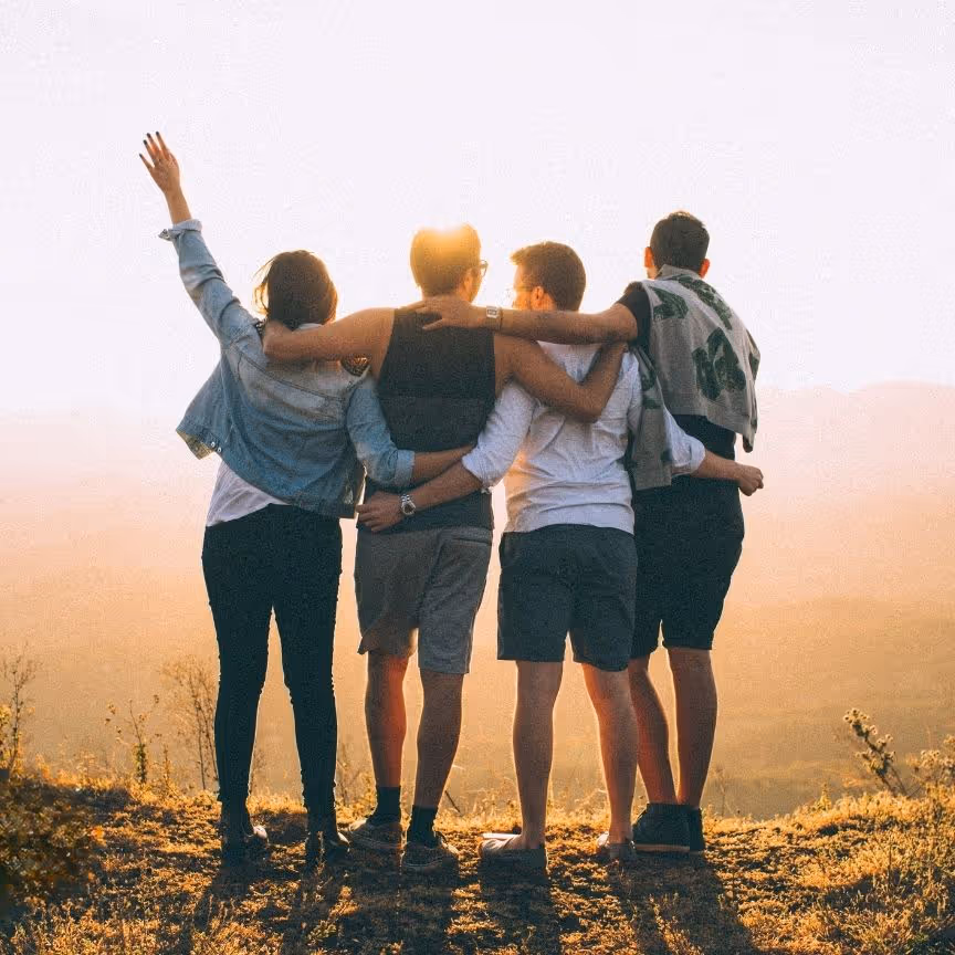Four friends with arms around each other, facing a sunset view on a mountaintop.