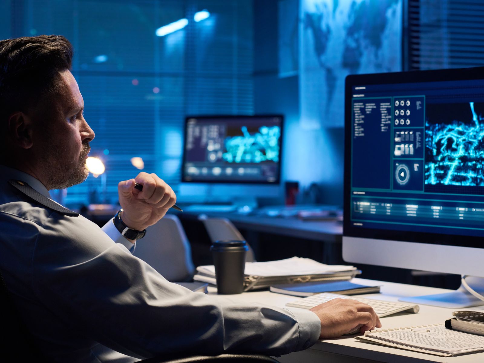 Man in uniform working late at desk with dual monitors displaying data and maps in dark office.