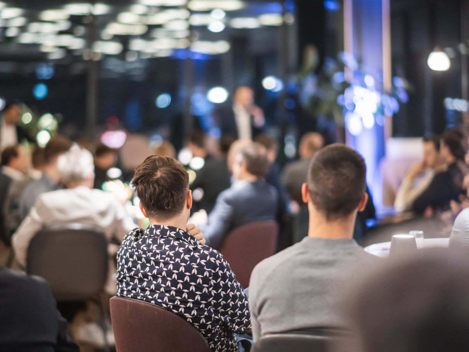 People seated indoors facing a speaker in a blurred background during a formal event or meeting.