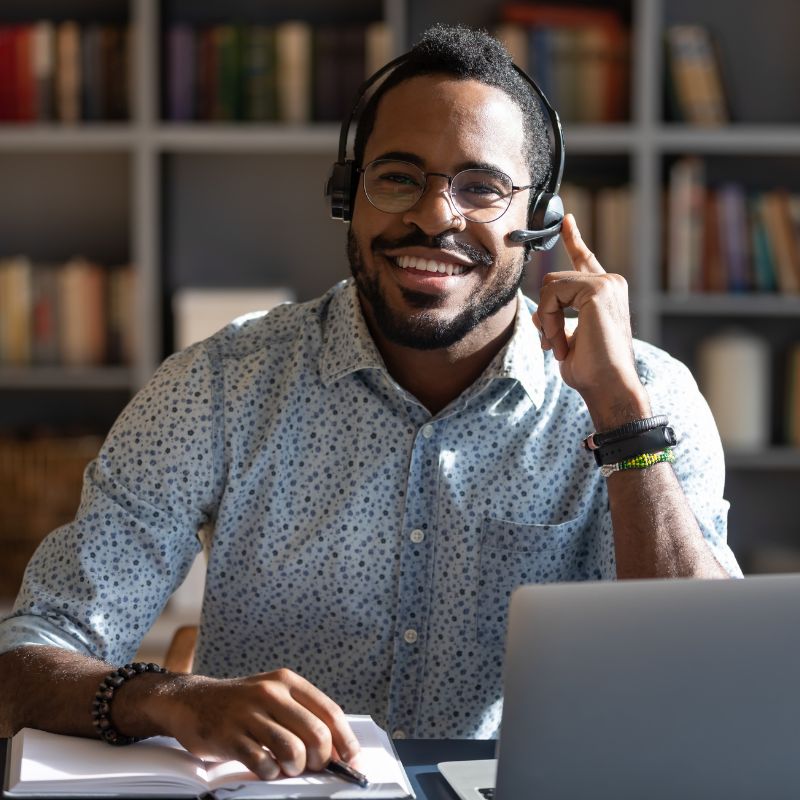 Smiling man with headset and glasses working at a desk with a laptop and notebook.