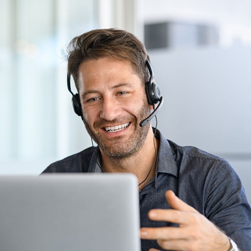 Smiling man wearing headset talking and gesturing while looking at a laptop screen.