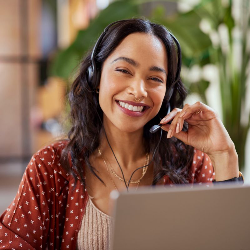 Smiling woman wearing headset and speaking into microphone while using laptop in a bright room.