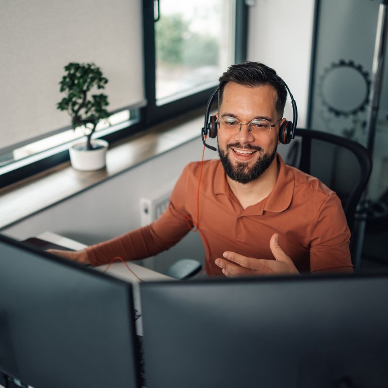 Smiling man with headset working at dual monitors in a modern office with a window and plant.