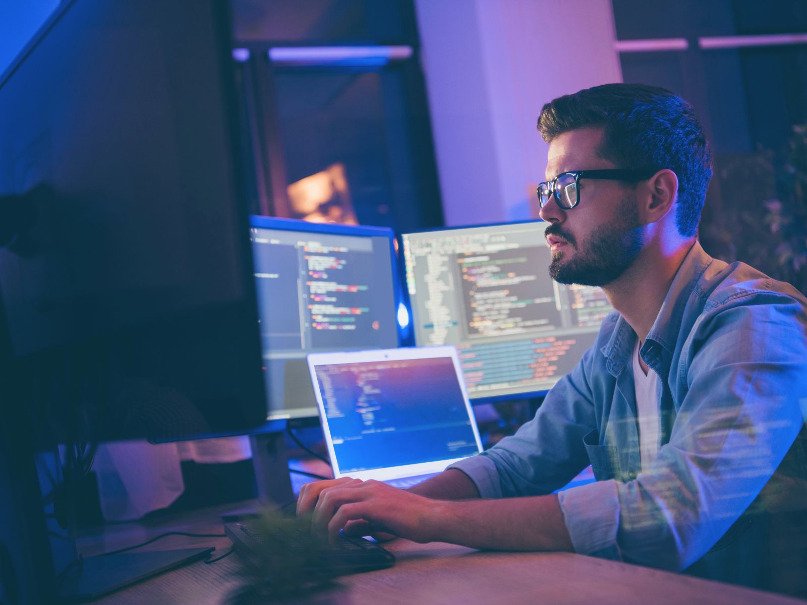 Man with glasses working on multiple screens displaying code in a dimly lit office.