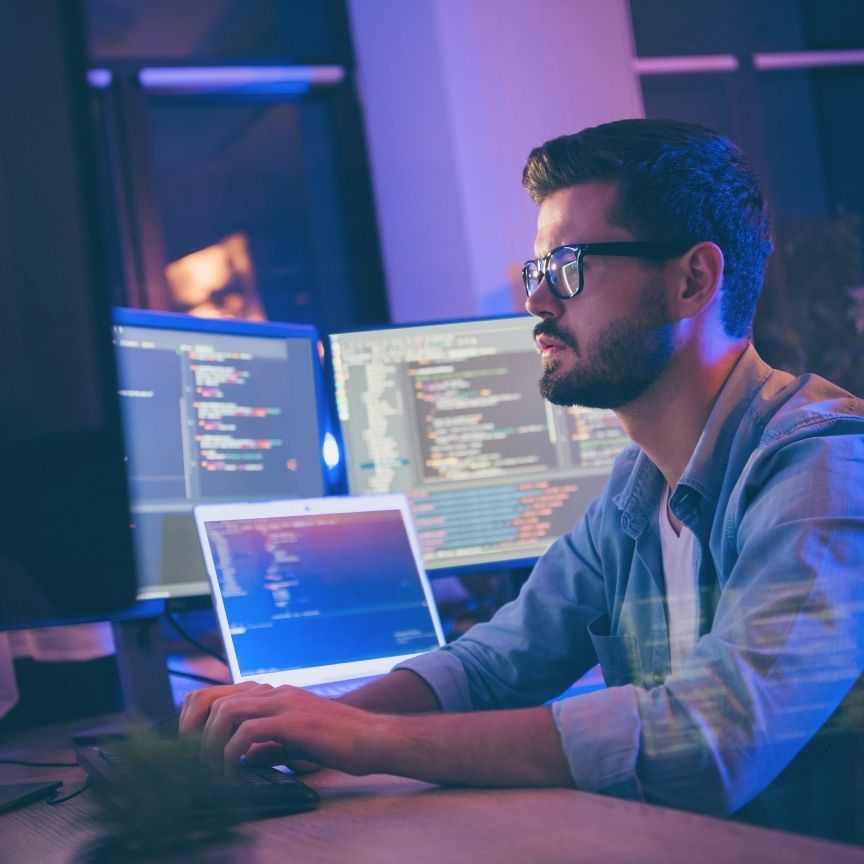 Man with glasses working on multiple screens displaying code in a dimly lit office.