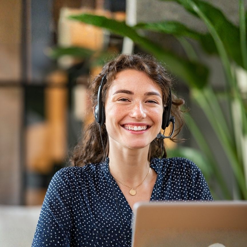 Smiling woman with headset working on a laptop in a modern office setting with plants.
