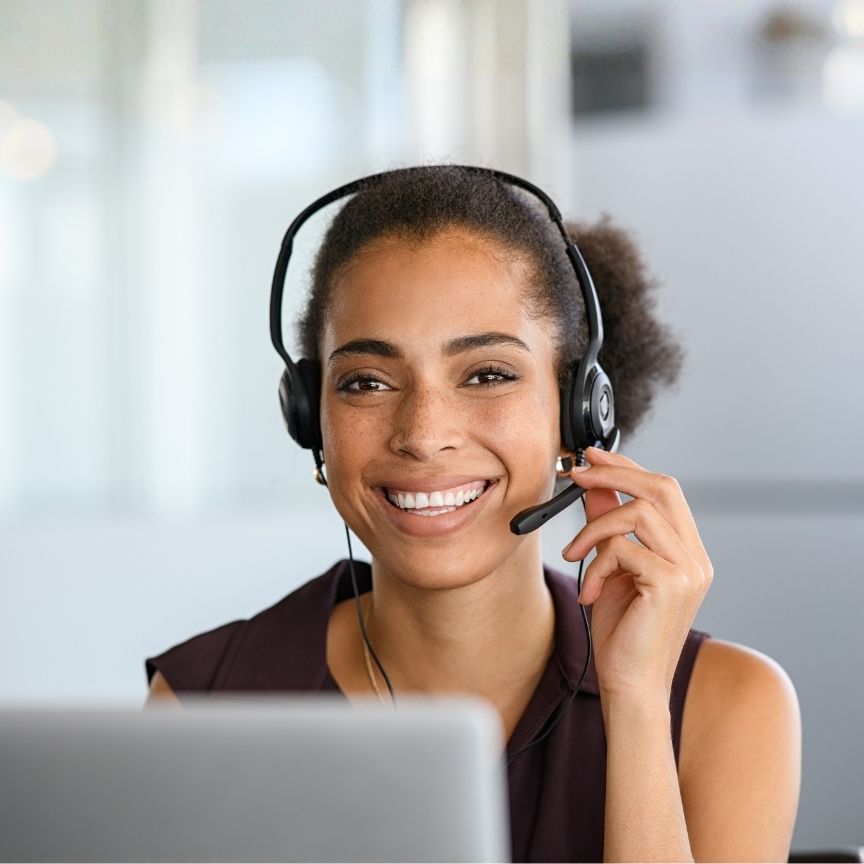Smiling woman wearing headset and speaking, sitting in front of a laptop in an office setting.