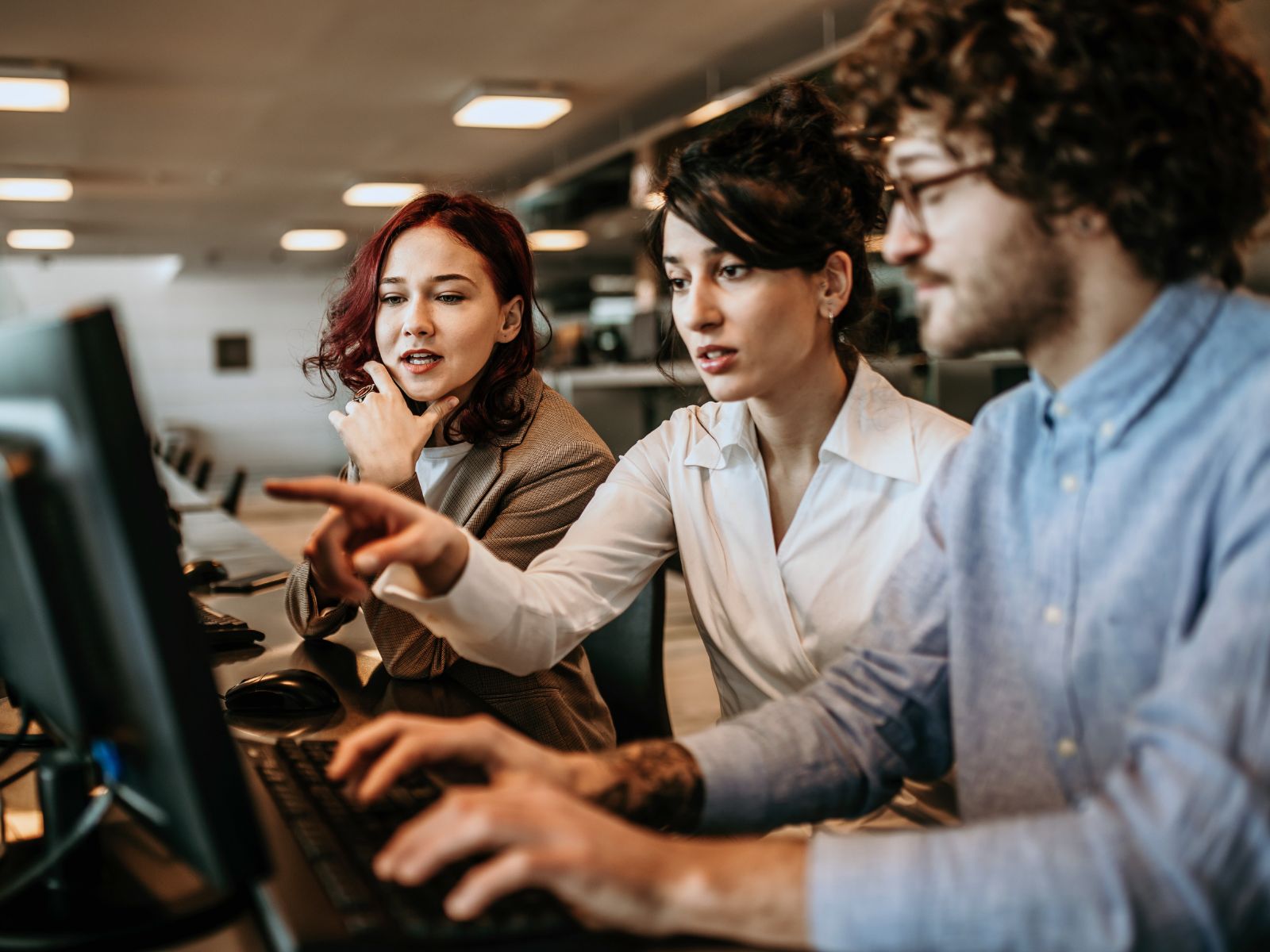 Three coworkers focused on a computer screen in a modern office, one pointing at the monitor.