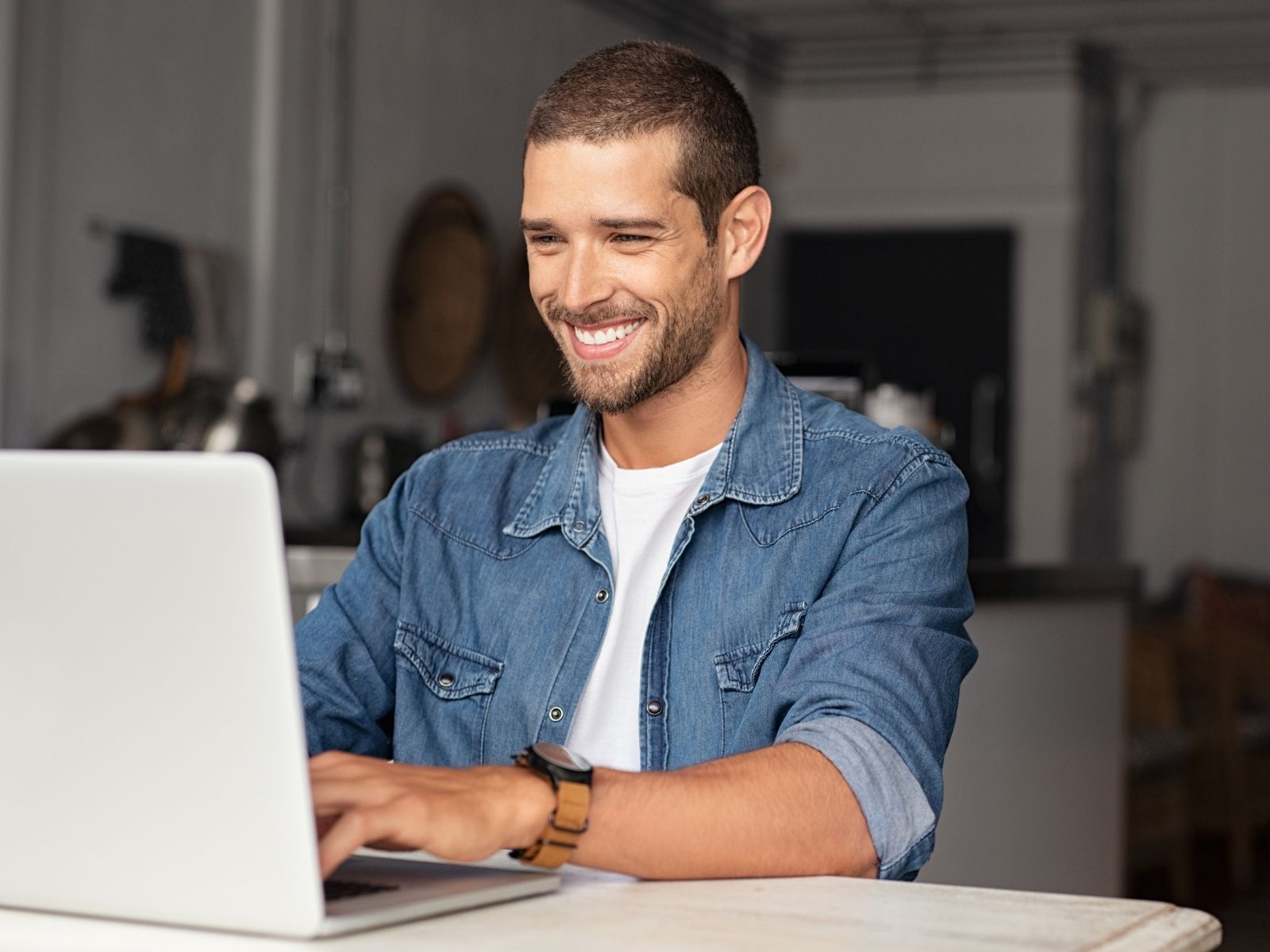 Smiling man in denim shirt using a laptop at a table indoors.