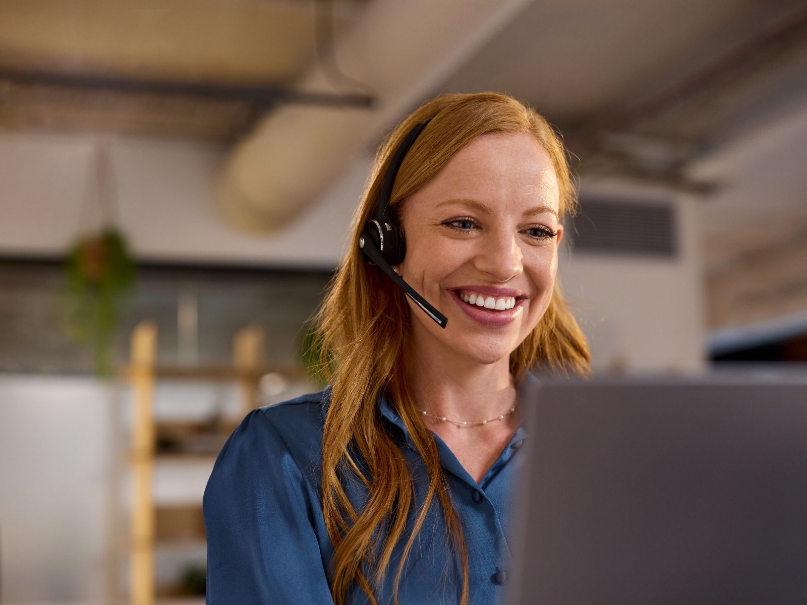 Smiling woman with red hair wearing headset looking at laptop in office setting.