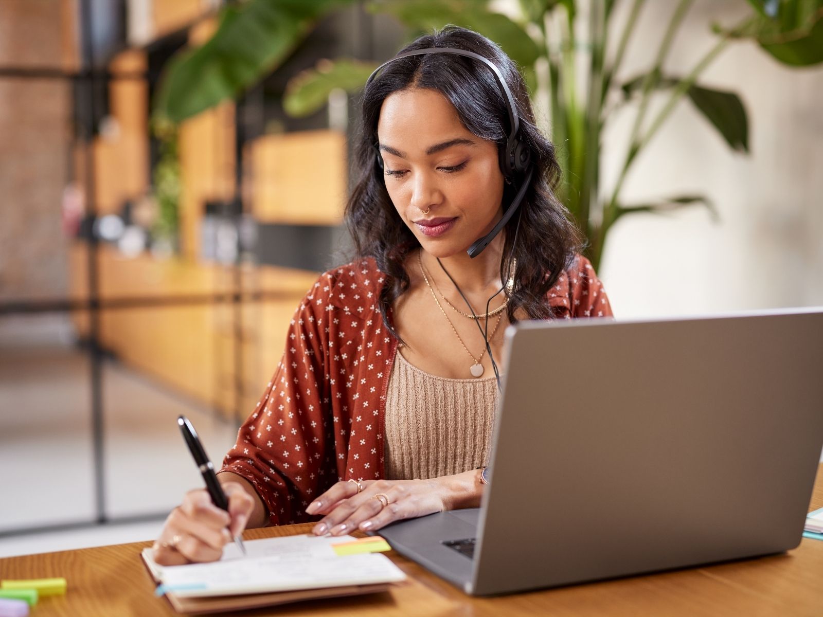 Woman wearing headset writing notes at a desk with an open laptop in a bright office.
